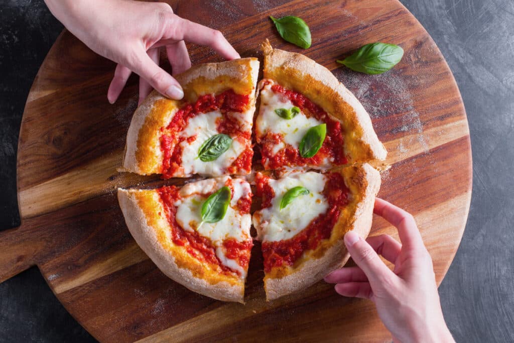 A top view of four slices of a margehrita veg pizza on a board with two people taking the slices