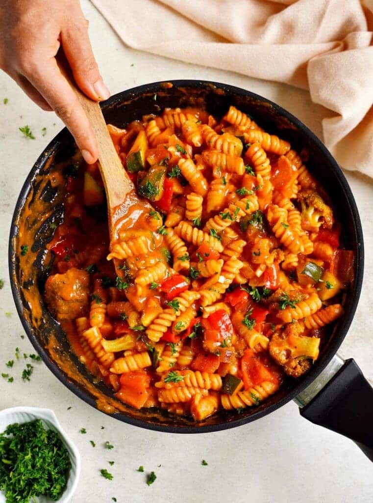 A top view of mixing pasta with veggies topped with cilantro in a pan with a ladle