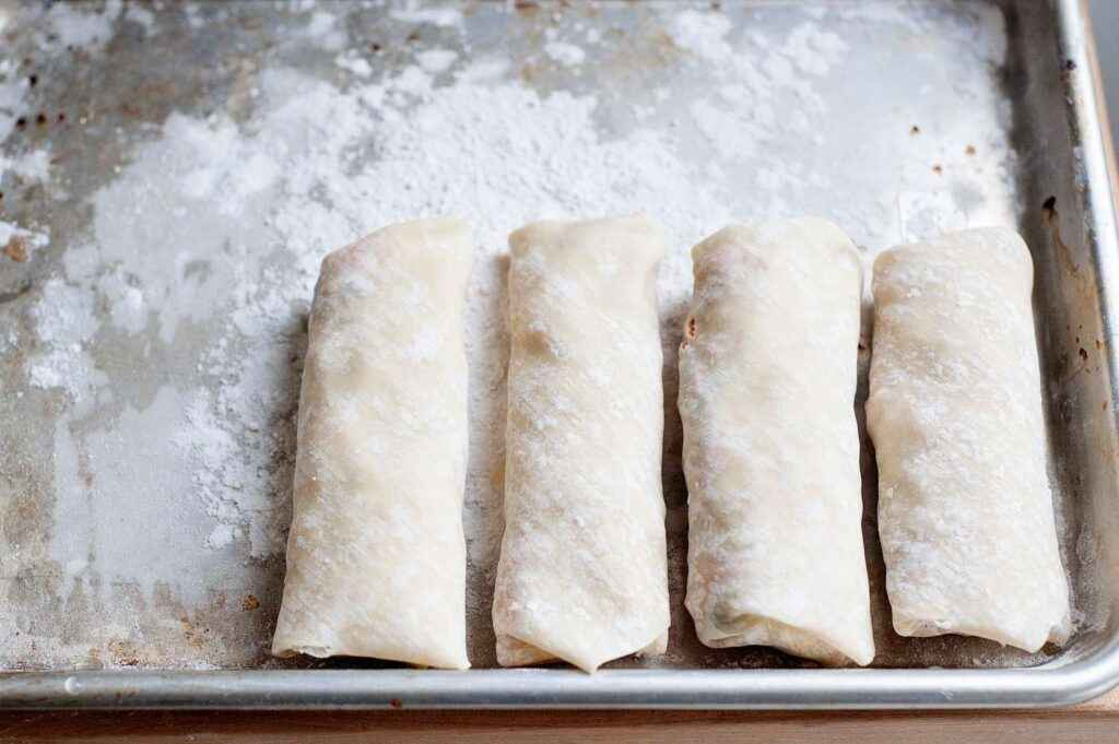 A view of raw egg rolls coated in flour on a baking tray ready to be cooked