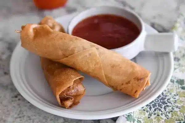 A view of two egg rolls in a white china dish along with tomato ketchup