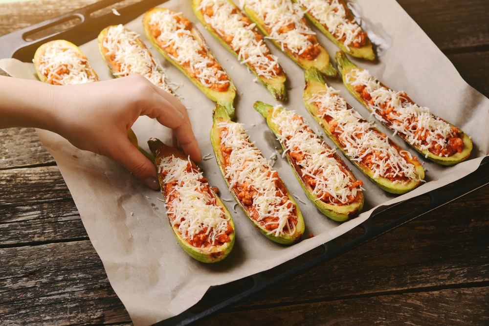 A view of a person putting zucchinis topped with cheese on a baking tray