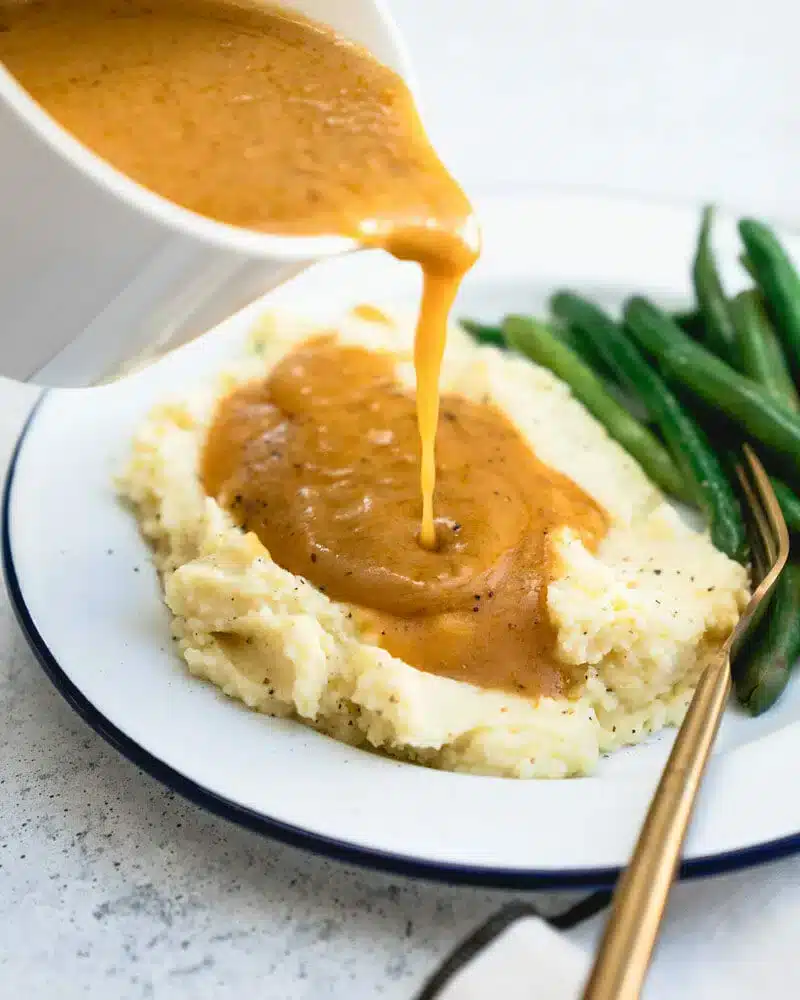 A top view of some vegetarian gravy being poured onto some mashed potatoes with a side of chillis