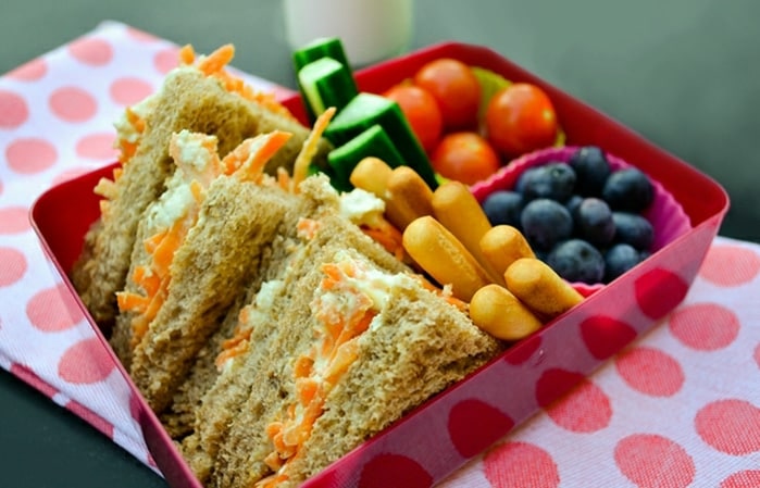 A view of a veggie sandwich served in a lunchbox along with some sweet fruits