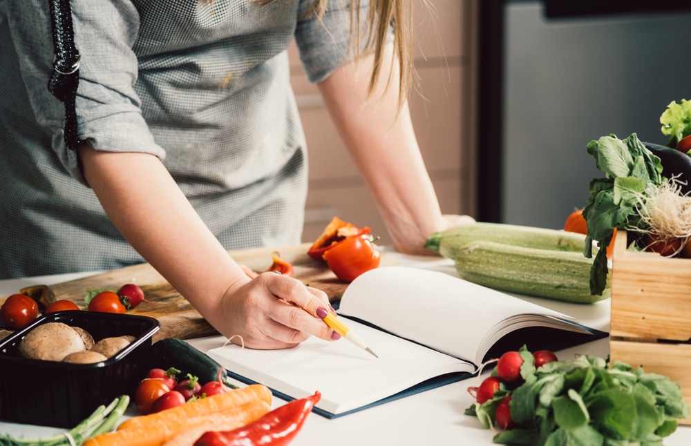 A view of a lady writing inside a cookbook with veggies around